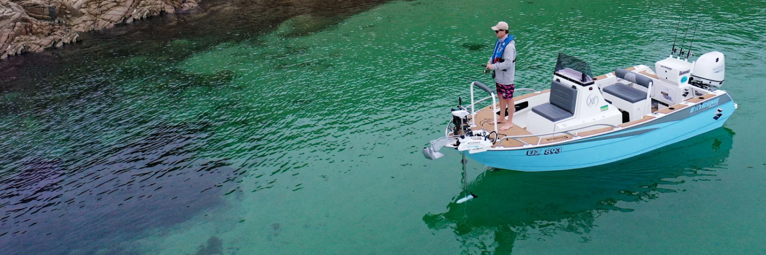 Pescador en una embarcación azul utilizando un motor eléctrico de proa Watersnake Geo-Spot GPS desplegado para mantener la posición en aguas cristalinas junto a la costa rocosa.