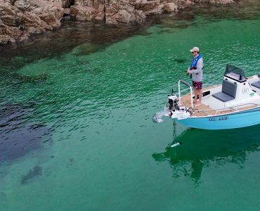 Pescador en una embarcación azul utilizando un motor eléctrico de proa Watersnake Geo-Spot GPS desplegado para mantener la posición en aguas cristalinas junto a la costa rocosa.