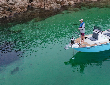 Pescador en una embarcación azul utilizando un motor eléctrico de proa Watersnake Geo-Spot GPS desplegado para mantener la posición en aguas cristalinas junto a la costa rocosa.