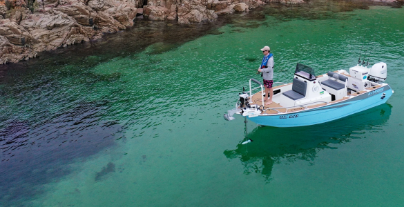 Pescador en una embarcación azul utilizando un motor eléctrico de proa Watersnake Geo-Spot GPS desplegado para mantener la posición en aguas cristalinas junto a la costa rocosa.
