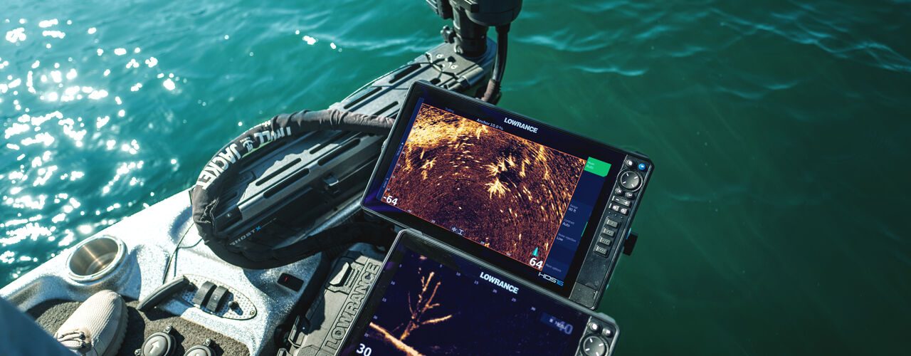 Vista desde la proa de una embarcación de pesca mostrando dos pantallas Lowrance operando en tiempo real con el sonar ActiveTarget 2 XL. En las pantallas se aprecian vistas detalladas del fondo y un árbol sumergido, junto al motor eléctrico desplegado en el agua.