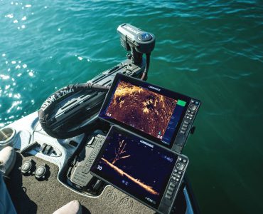 Vista desde la proa de una embarcación de pesca mostrando dos pantallas Lowrance operando en tiempo real con el sonar ActiveTarget 2 XL. En las pantallas se aprecian vistas detalladas del fondo y un árbol sumergido, junto al motor eléctrico desplegado en el agua.