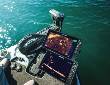 Vista desde la proa de una embarcación de pesca mostrando dos pantallas Lowrance operando en tiempo real con el sonar ActiveTarget 2 XL. En las pantallas se aprecian vistas detalladas del fondo y un árbol sumergido, junto al motor eléctrico desplegado en el agua.
